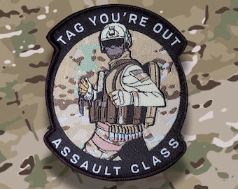 May include: A circular patch featuring a soldier in combat gear, with the text "TAG YOU'RE OUT ASSAULT CLASS" in a black border. The soldier is depicted in tan and brown tones, set against a camouflage background.