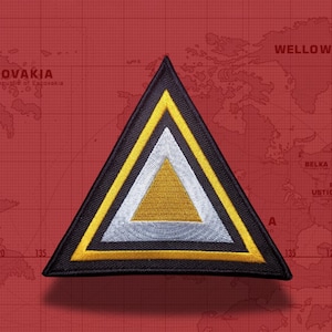 May include: A triangular embroidered patch featuring a black outer border, a thick yellow middle border, and a silver-grey inner border surrounding a solid gold-coloured centre triangle. The patch is displayed against a red map background.