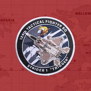 May include: A round, black and white patch with a white border. The patch features a gray and brown fighter jet flying over a white cloud. The text "124th Tactical Fighter SQ" is written in a white arc around the jet. Below the jet, the text "Strider 1 "Trigger" is written in a white arc.