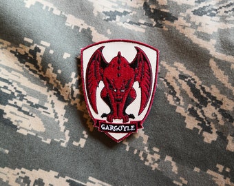 May include: Red embroidered patch featuring a gargoyle design on a white background. The patch is shaped like a shield and has the word "GARGOYLE" written below the gargoyle.