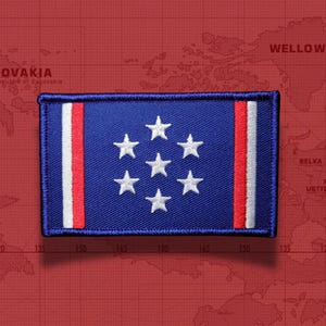 May include: A rectangular embroidered patch featuring a blue field with seven white stars arranged in a circular pattern. The patch includes vertical white and red stripes on both the left and right sides, finished with a dark blue stitched border.