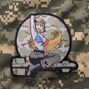 Könnte beinhalten: Ein runder bestickter Aufnäher mit einer Anime-Figur mit langem blonden Haar, die einen Helm, ein Bikini-Oberteil mit blau-weißem Sternenmuster und eine dunkle Hose trägt und auf einer Rakete vor einem Tarnhintergrund sitzt.