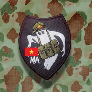 May include: A shield-shaped embroidered patch with a black background featuring a white ghost wearing a green helmet with a red emblem and carrying three olive green ammunition pouches. The patch includes a small rectangular flag of Vietnam.