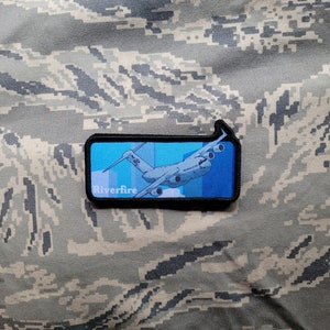 Peut inclure: Un patch bleu et blanc avec un avion militaire et le texte "Riverfire" sur un fond camouflage.