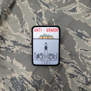 May include: A black and white patch with the text "ANTI-ARMOR" in red. The patch depicts a military aircraft dropping bombs on a tank.