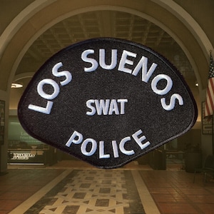 Puede incluir: Parche negro con texto blanco que dice "LOS SUENOS SWAT POLICE". El parche tiene una parte superior curvada y una superficie texturizada. El fondo muestra el interior de un edificio con una bandera americana.