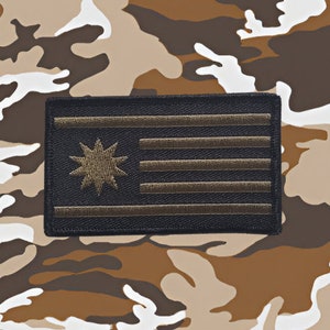 以下が含まれることがあります： 星形のバーストとストライプが施された黒と茶色の刺繍パッチ。パッチは迷彩柄の背景の上にあります。