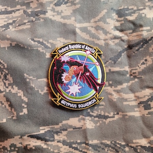 Könnte beinhalten: Ein gestickter Aufnäher in Schwarz, Rot und Weiß mit einem stilisierten Greifvogel im Flug. Der Aufnäher ist von einem gelben Kreis mit dem Text "Föderative Republik Aurelia Gryphus Geschwader" umgeben.