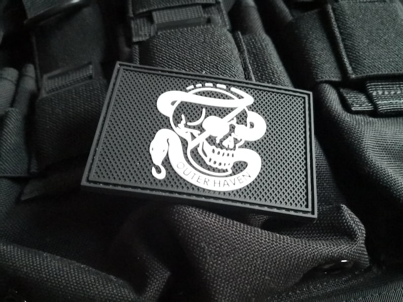 Op de afbeelding: Witte patch met een schedel en slang ontwerp. De tekst "Outer Haven" staat onder de schedel. De patch is op een zwarte achtergrond.
