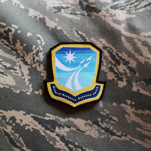 May include: A blue, yellow, and white embroidered patch with the text "General Resource Defence Force" around the edge. The patch features a white star and three white missiles on a blue and white background.