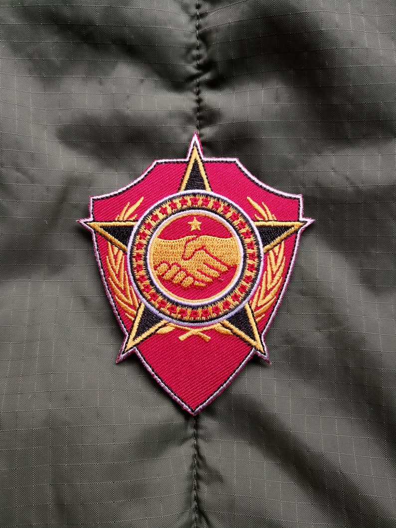 May include: An embroidered patch in red, white and gold, featuring a star, a handshake and a wreath. The patch is shield-shaped with a black outline.