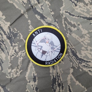 May include: A black and yellow circular patch with the text "ANTI RADIATION" around the edge. The centre of the patch features a white-haired anime-style character wearing a white shirt and black shorts.