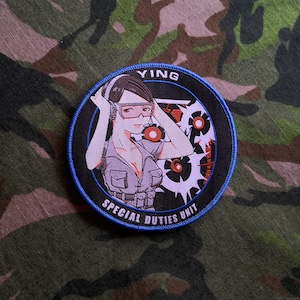 Könnte beinhalten: Ein gestickter schwarzer und weißer Aufnäher mit einer Comic-Frau, die Kopfhörer und eine Militäruniform trägt. Der Aufnäher hat einen blauen Rand und den Text "Ying Special Duties Unit".