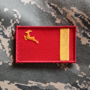 May include: Red and gold embroidered patch with a stylized deer and a vertical gold stripe.