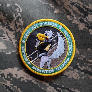 May include: A green and yellow embroidered patch with a cartoon bird holding a gun. The patch reads "22nd Air Division 4th Tactical Fighter Squadron".