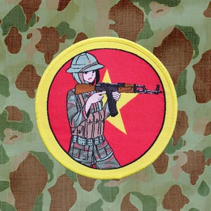 May include: A yellow and red embroidered patch featuring a cartoon woman in a green and brown camouflage uniform holding a rifle. The woman is wearing a green helmet and a red star is in the background.