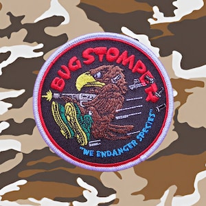 May include: A black and red embroidered patch with a cartoon eagle holding a gun and the text "Bug Stomper" and "We Endanger Species".