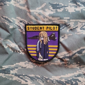 Peut inclure: Un écusson brodé violet et jaune avec une illustration de dessin animé d'une femme portant une veste bleue et tenant un livre. Le texte "STUDENT PILOT" est en haut de l'écusson.