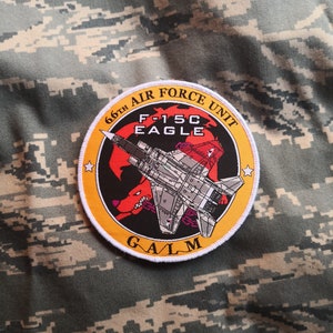 Può includere: Una toppa gialla e bianca con il testo "66th Air Force Unit F-15C Eagle GALM" e un'immagine di un aereo da combattimento con un lupo rosso e nero.