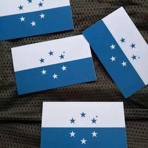 May include: Three white and blue rectangular flags with a blue stripe and white stars. The flags are on a brown textured surface.
