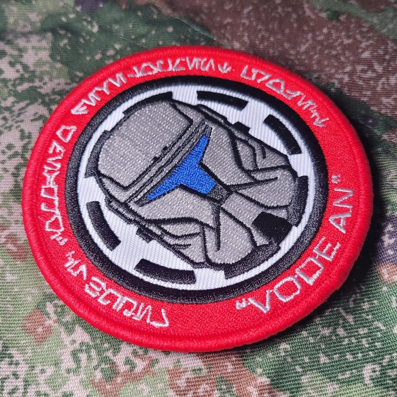 Pu&ograve; includere: Una toppa rotonda con bordo rosso, raffigurante un design di elmetto grigio con accenti blu. La toppa presenta dettagli bianchi e neri e testo in un motivo circolare. Il testo include le parole "VODE AN".