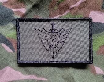 May include: A black and green embroidered patch with a stylized eagle design and a sword. The patch is surrounded by a black border.