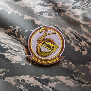 May include: A brown and white embroidered patch with a yellow snake design. The text "GRABBER" is on the snake. The text "229th TACTICAL FIGHTER SQ" is on the bottom of the patch.