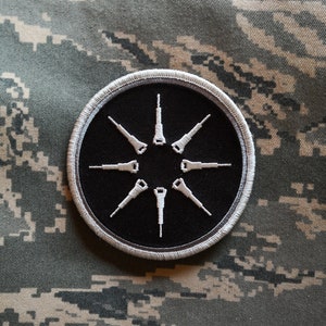 May include: A black and white embroidered patch with a circle design. The circle has a black background and a white outline. Inside the circle are eight white stylized arrows pointing outwards from the center.