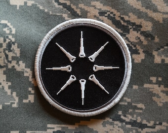 May include: A black and white embroidered patch with a circular design. The circle has a black background and a white outline. Inside the circle are eight white stylised arrows pointing outwards from the centre.