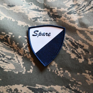 Könnte beinhalten: Ein weißer und blauer Stoff-Patch mit dem Wort "Spare" in weiß auf weißem Hintergrund gestickt. Der Patch ist schildförmig und hat einen blauen Rand.