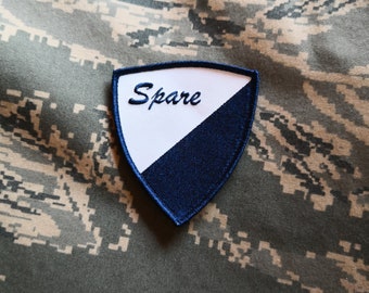 May include: A white and blue fabric patch with the word "Spare" embroidered in white on a white background. The patch is shaped like a shield and has a blue border.