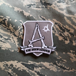 May include: A white and grey embroidered patch with a stylised bone arrow design and the text "BONE ARROW SQUADRON".