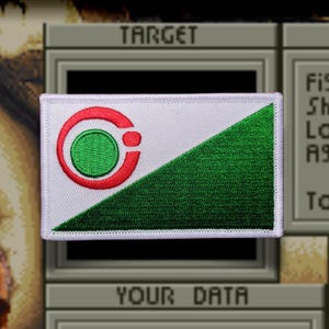 May include: A rectangular embroidered patch featuring a diagonal split design. The upper left section is white, and the lower right section is green. A red circular emblem with a green center is positioned on the white portion of the patch.