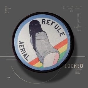 May include: A circular embroidered patch featuring a person with long dark hair wearing a gray hoodie, positioned against a background with diagonal red, yellow, and green stripes. The text AERIAL REFULE is printed in black along the curved border.