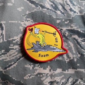 Może przedstawiać: Naszywka haftowana na żółto i czerwono z postacią z kreskówki jadącą na myśliwcu. Postać ma na sobie czerwony kapelusz i trzyma mikrofon. Tekst na naszywce brzmi "I am Saam".