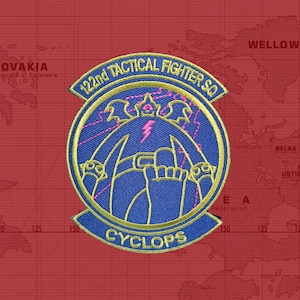 Peut inclure: Un écusson brodé doré sur fond bleu. L'écusson représente un cyclope stylisé avec un éclair et le texte "122nd Tactical Fighter Sq Cyclops".
