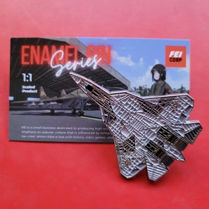 May include: A silver-colored enamel pin of a fighter jet with a detailed design. The pin is on a red background with a card that says "Enamel Pin Series" and "1:1 Scaled Product".