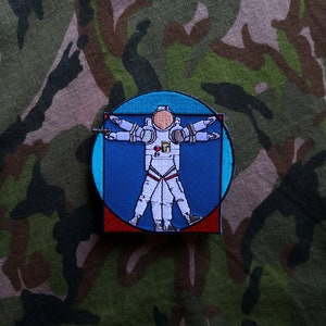 May include: An embroidered patch featuring a white astronaut with outstretched arms, resembling the famous Vitruvian Man, inside a blue circle with a red and white frame.