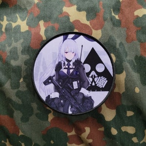 May include: A round, white and black patch featuring a cartoon image of a woman with long, white hair holding a rifle. The woman is wearing a black outfit with a white collar and a black tie. The patch has a black triangle with a skull and crossbones in the centre. The text "E.F.Y" is printed below the triangle.