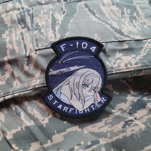 Peut inclure: Un écusson brodé bleu et blanc avec une fille de dessin animé anime dormant sur un nuage. L'écusson porte l'inscription "F-104 STARFIGHTER" sur le bord.