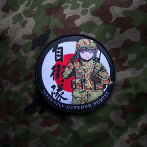 May include: A black and white circular patch with a red circle and Japanese text. The patch features a cartoon anime woman in a camouflage uniform saluting. The text reads "Japan Self-Defense Forces".