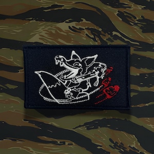 May include: A rectangular navy blue patch with a white embroidered design of a stylized creature holding a weapon, with red accents. The patch is set against a camouflage background.