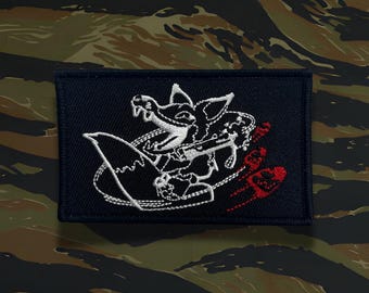 May include: A rectangular navy blue patch with a white embroidered design of a stylized creature holding a weapon, with red accents. The patch is set against a camouflage background.