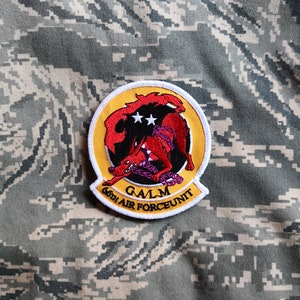 May include: A circular patch with a red and black wolf-like creature design, two white stars, and the text "GALM 66th AIR FORCE UNIT" in white. The patch has a yellow border and is set against a camouflage background.