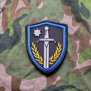 May include: A black and blue embroidered patch with a white sword and a gold wreath. The patch features a white star with a single point.