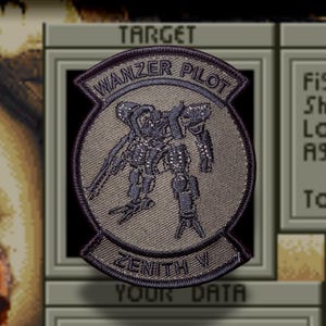 May include: A circular embroidered patch featuring a dark grey mecha robot in the centre. The top arc reads WANZER PILOT and the bottom arc reads ZENITH V in dark thread. The background of the patch is a textured grey with a dark border.