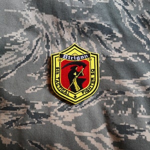 May include: A yellow and black embroidered patch with a skull and crossbones design. The patch reads "96th Tactical Fighter Squadron" and "Strigon".