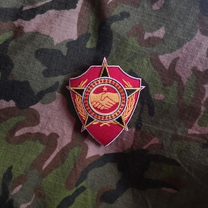 Könnte beinhalten: Ein gestickter Aufnäher in Rot und Gold mit einem Stern, einem Handschlag und einem Kranz. Der Aufnäher befindet sich auf einem Tarnstoff-Hintergrund.