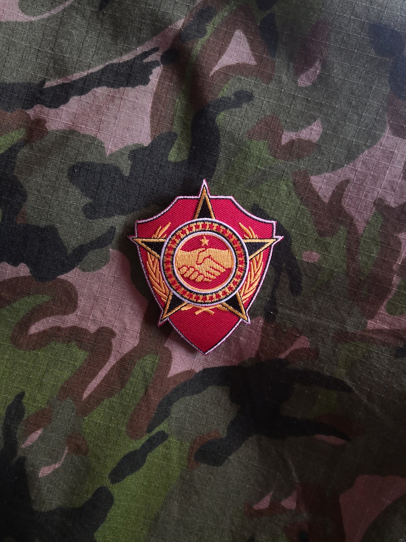 May include: A red and gold embroidered patch featuring a star, a handshake, and a wreath. The patch is displayed on a camouflage fabric background.