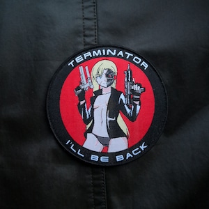 Może przedstawiać: Okrągła naszywka w kolorze czarnym i czerwonym z napisem "TERMINATOR I'LL BE BACK" w kolorze białym. Naszywka przedstawia kreskówkową postać kobiety z blond włosami trzymającej dwie pistolety. Twarz kobiety jest w połowie ludzka, a w połowie robotyczna.